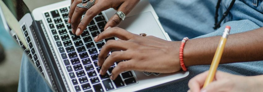 High angle crop faceless young multiracial women sitting in park with open laptop while writing with pencil in notepad